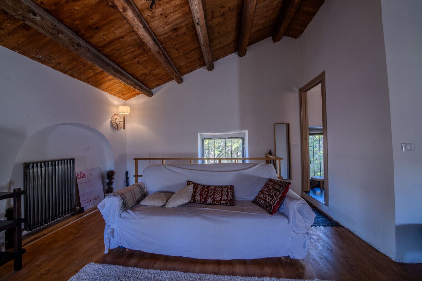 Ein Schlafzimmer mit Holzboden, einem Bett mit weißer Bettwäsche und gemusterten Kissen, einem Fenster und einer Heizung.