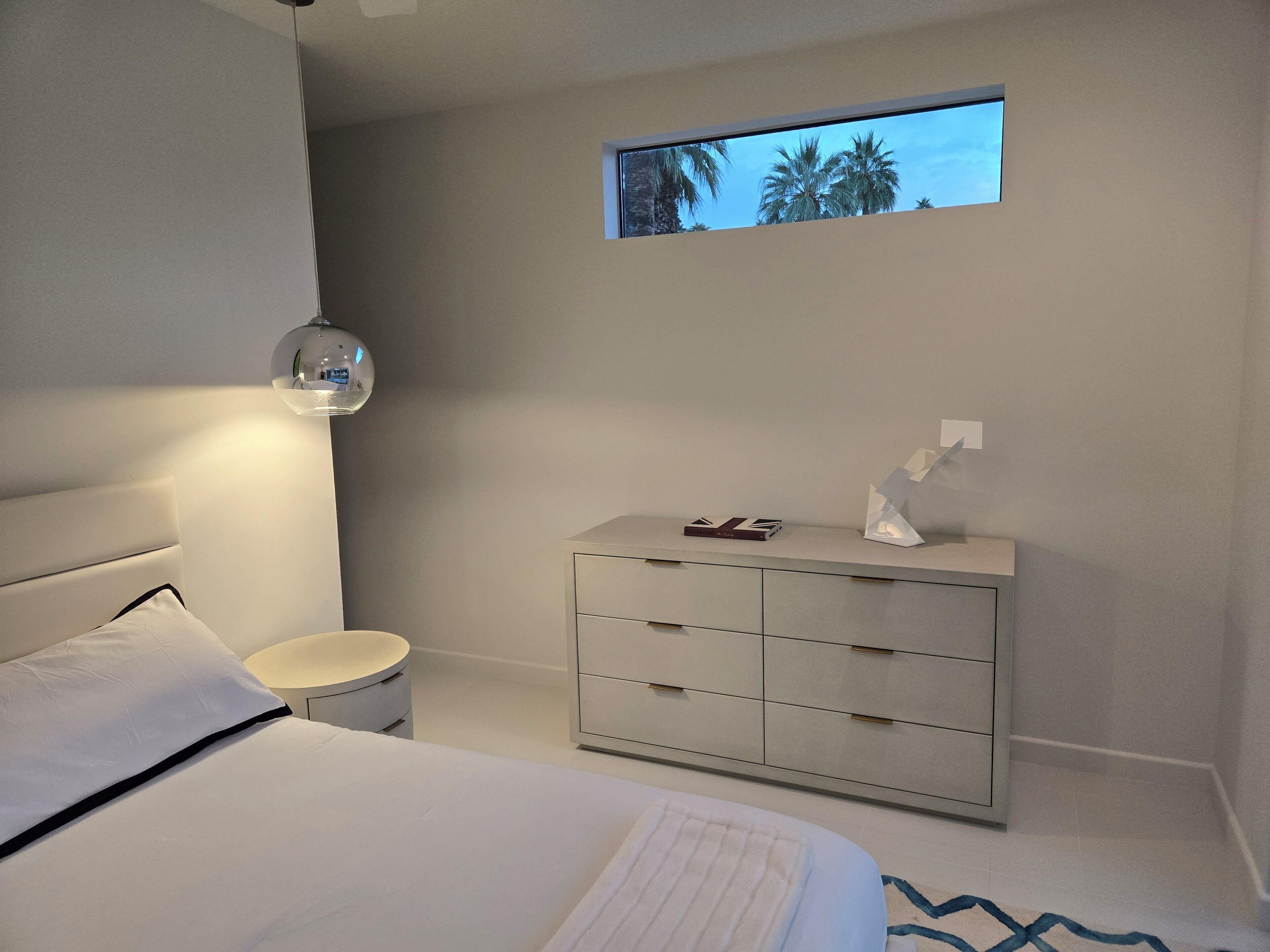 Ein Schlafzimmer mit Bett, Nachttischlampe, Nachttisch, Kommode und einem kleinen Fenster mit Blick auf Palmen.