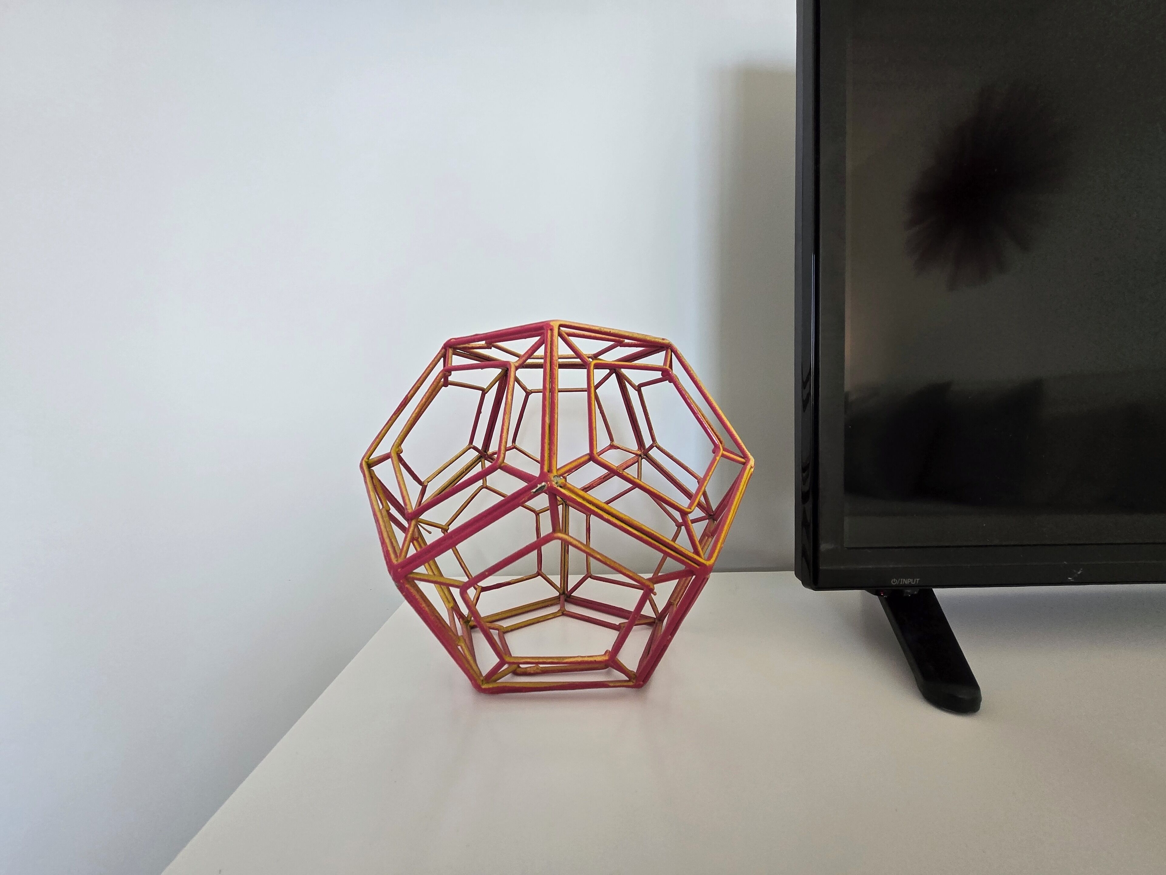 Uma estrutura geométrica, semelhante a uma colmeia, com moldura metálica, colocada sobre uma superfície branca ao lado de um objeto preto, possivelmente uma televisão.