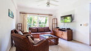 Een woonkamer met rieten meubels, een plafondventilator, een televisie en een balkon met gordijnen.