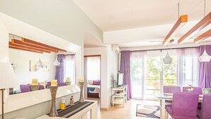 A modern living room with a sofa, a dining table, and a TV. There is a balcony with a view of trees. - Infiniti Blu, Family Suite, Private Beach! (Sosúa)