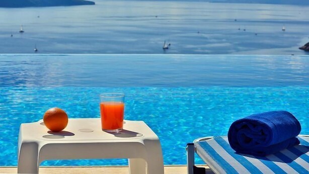 Blick über den Pool mit blauem Wasser, ein Glas Orangensaft und ein zusammengerolltes Handtuch auf einem Stuhl.