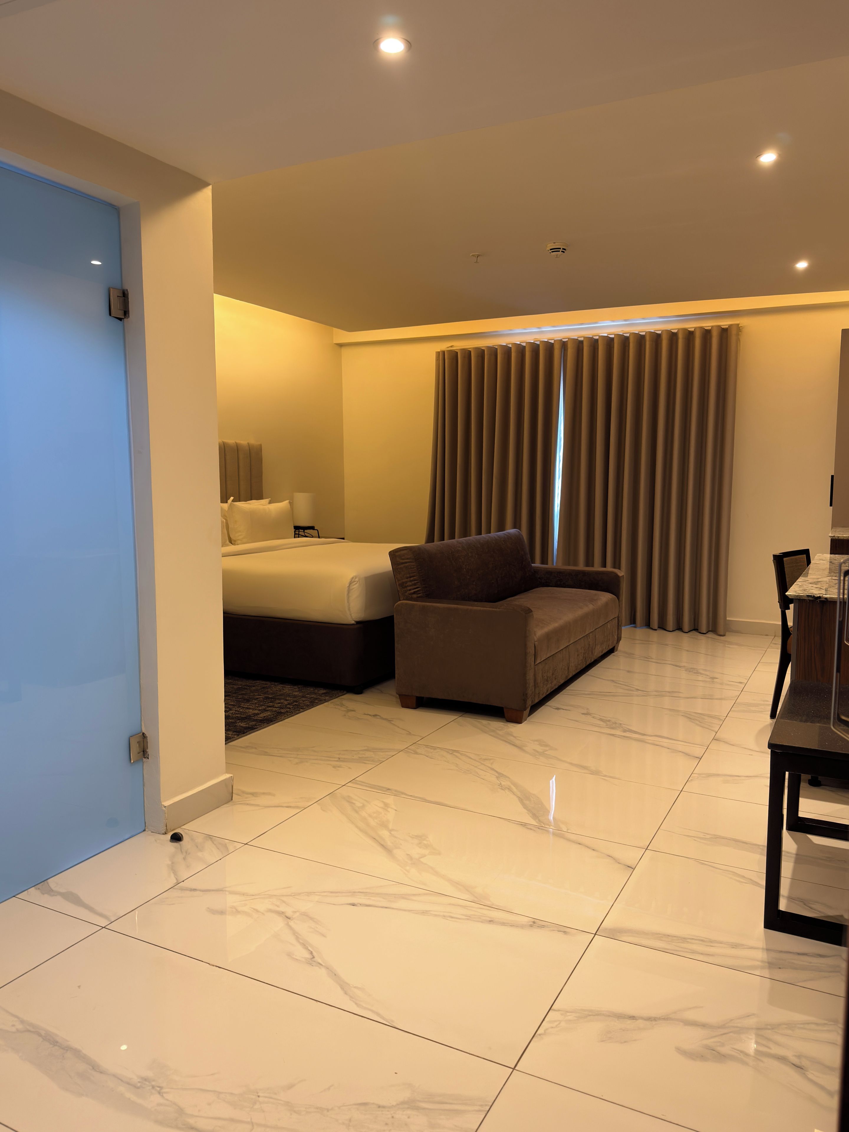 Quarto de hotel com cama, sofá, mesa de jantar e uma janela grande com cortinas.