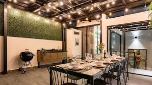 Een eetgedeelte met een lange tafel gedekt voor zes personen, omringd door stoelen. Een grill is links zichtbaar, en er is een muur met een groen, reliëf oppervlak achter de tafel.