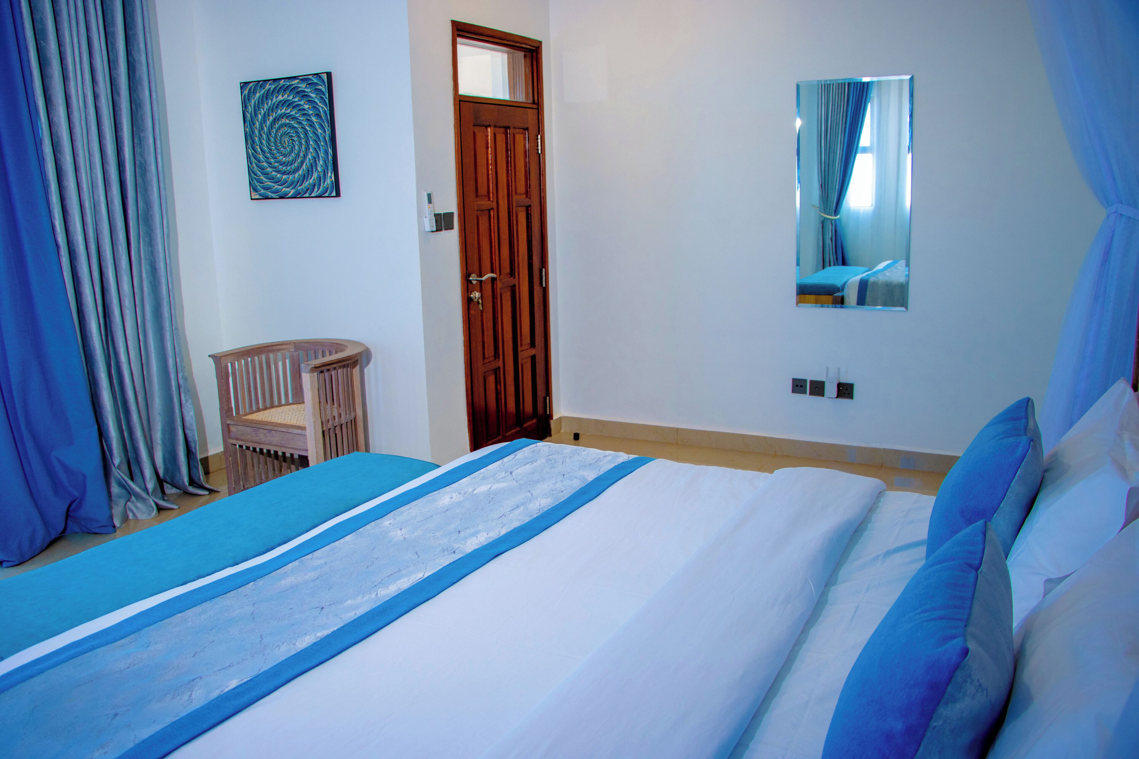 Une chambre à coucher comprenant un lit, une porte en bois, une chaise, un miroir et un mur orné d’une œuvre d’art circulaire.