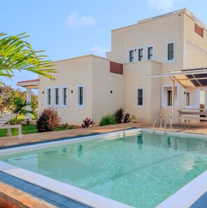 A swimming pool with a clear blue water surface, surrounded by a building with multiple windows and a covered seating area. - 4-bedroom villa in enchanting Pazuri in Kilifi County with AC, WiFi (Kilifi County)