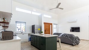 Et moderne køkken med hvide skabe, en central ø og en indbygget ovn. Der er et spiseområde med en sofa og et fjernsyn ophængt på væggen.