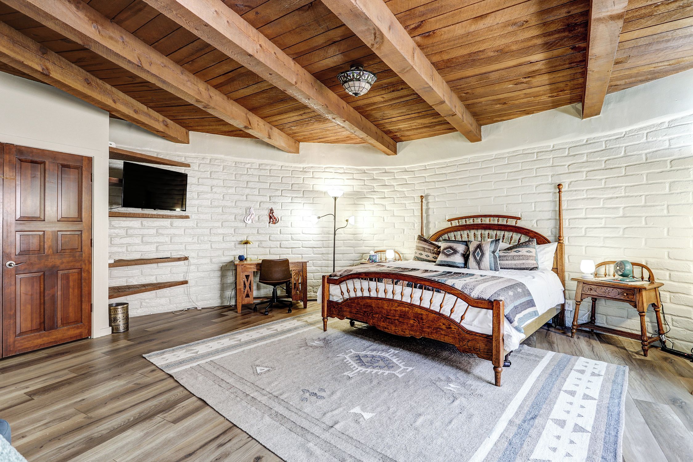 A bedroom with a wooden ceiling, a brick wall, a bed with a wooden frame, a desk, a chair, and a television mounted on the wall.