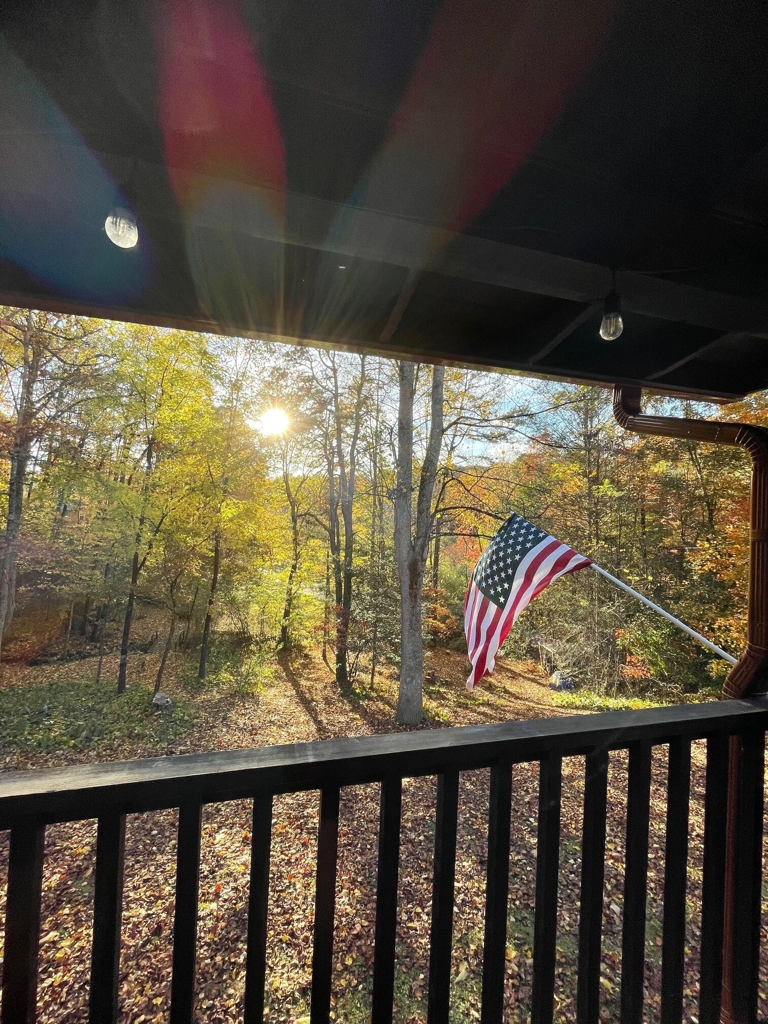 A view from a covered porch with an American flag, trees, and sunlight.