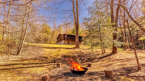 Una cabaña en un bosque con una hoguera al frente.