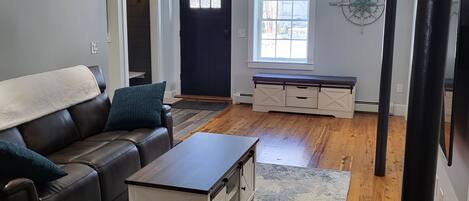 A living room with a leather sofa, a coffee table, and a white cabinet. There is a window with blinds, a door, and a wall clock.