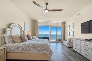 Une chambre à coucher comprenant un lit, un ventilateur de plafond, une télévision, une commode et un balcon avec vue sur l’océan.