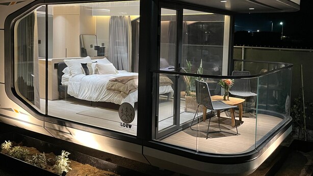 A modern, glass-enclosed bedroom with a bed, curtains, and a small table with a chair.