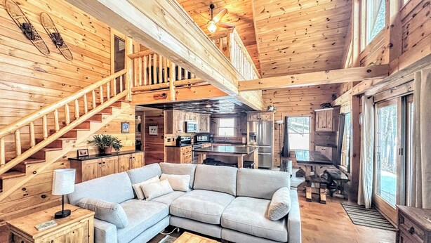 Un salón espacioso con una escalera de madera, un amplio sofá en forma de L y una cocina al fondo.