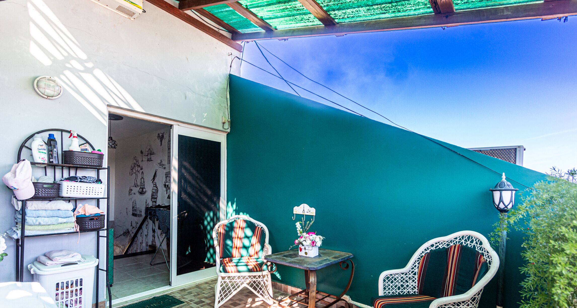 Un patio couvert agrémenté d’un mur végétal, de fauteuils en osier, d’une petite table avec une lampe et d’un panier à linge.