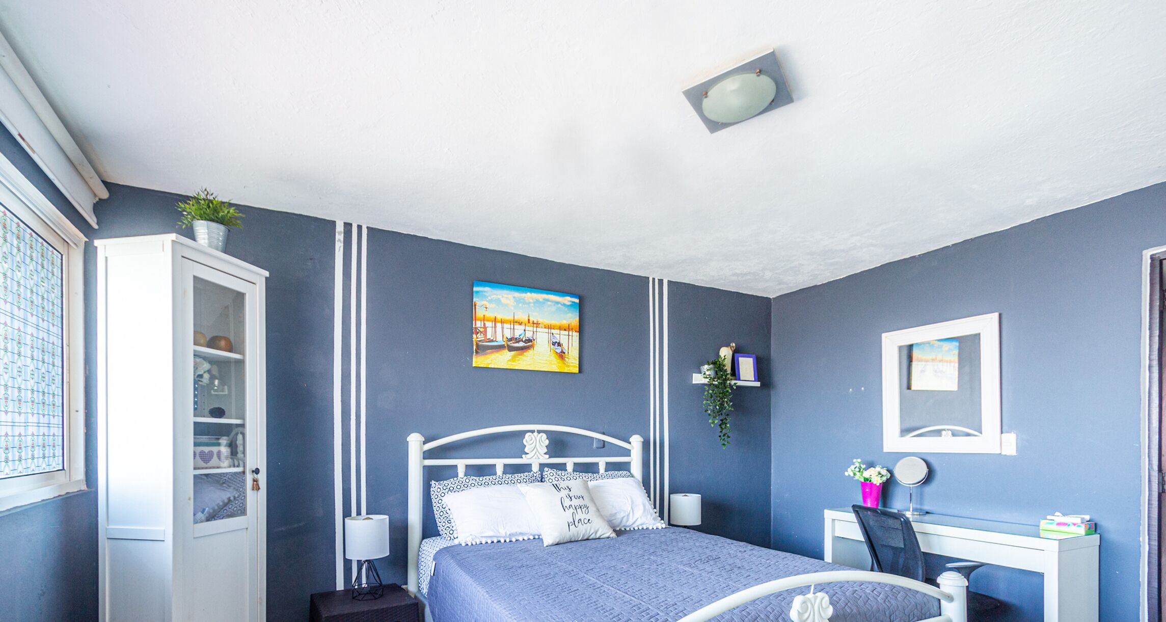 Een slaapkamer met een bed, een bureau, een stoel, een raam en een plank met spullen.
