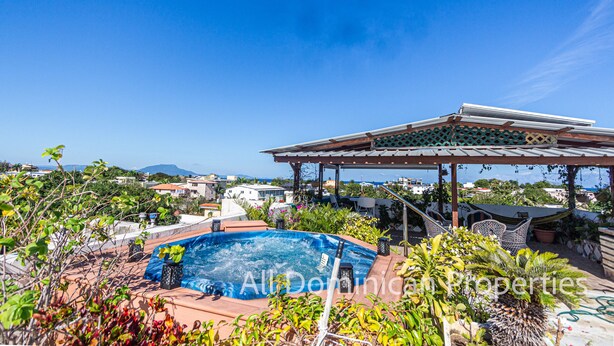 Una piscina al terrat amb una pèrgola i mobles d'exterior, envoltada de vegetació exuberant i amb vista a una vila costanera.