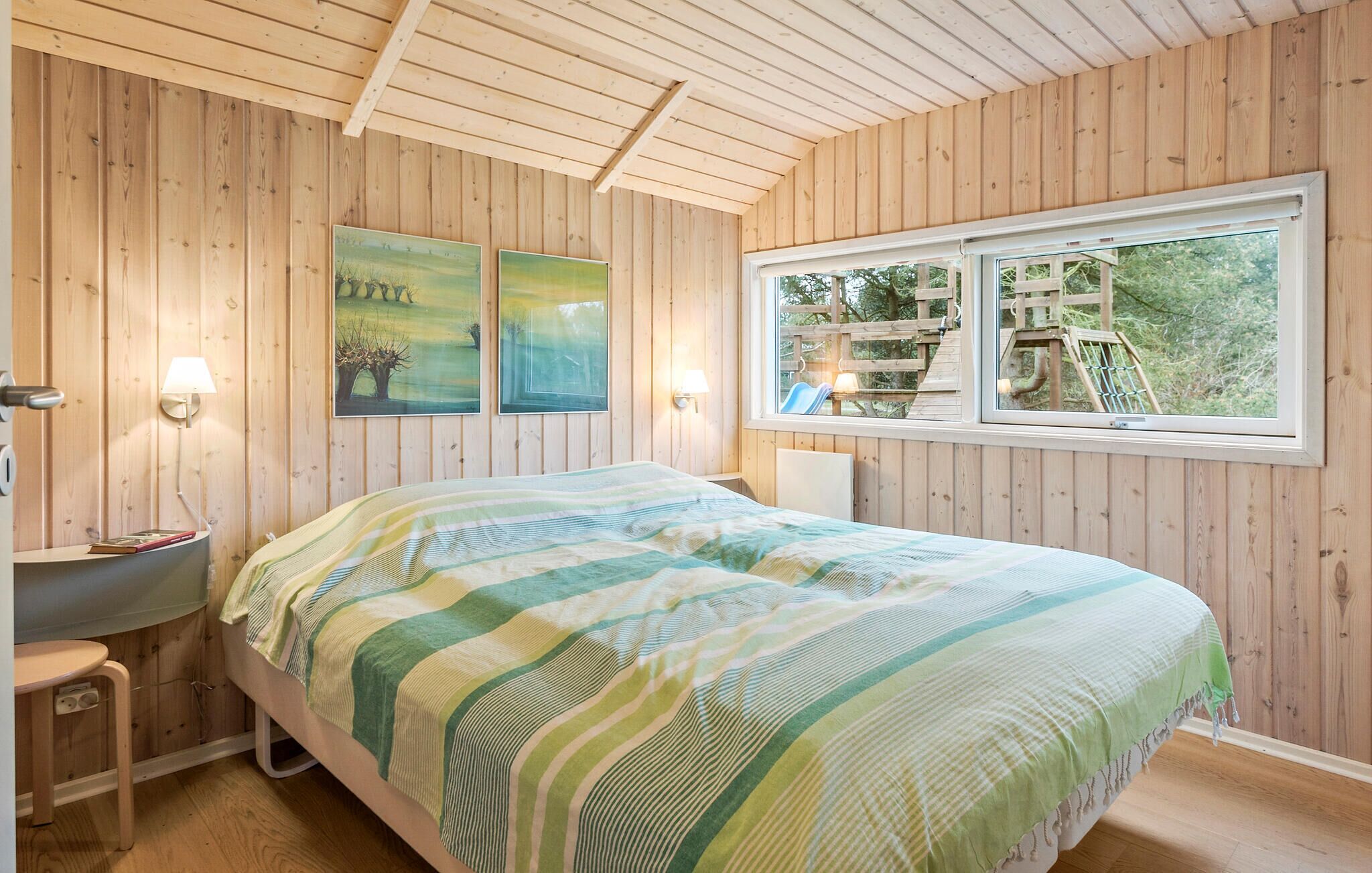 A wooden-paneled bedroom with a bed, a bedside table, two wall-mounted lamps, and a window with a view of a wooden deck and trees.