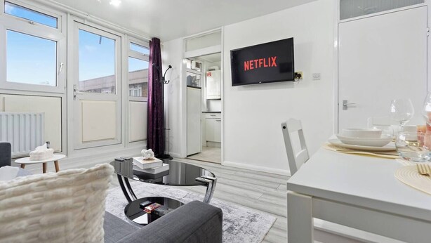 A modern living room with a sofa, coffee table, and a dining area with a table set for two. A flat-screen TV mounted on the wall displays "NETFLIX".