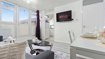 Un salon moderne avec un canapé, une table basse et un coin repas avec une table dressée pour deux personnes. Un téléviseur à écran plat, fixé au mur, affiche le logo « NETFLIX ».
