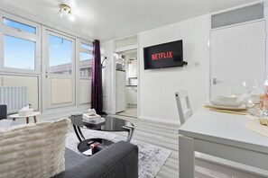 Una sala de estar moderna con un sofá, una mesa de centro y un comedor con una mesa dispuesta para dos. Un televisor de pantalla plana montado en la pared muestra “NETFLIX”.