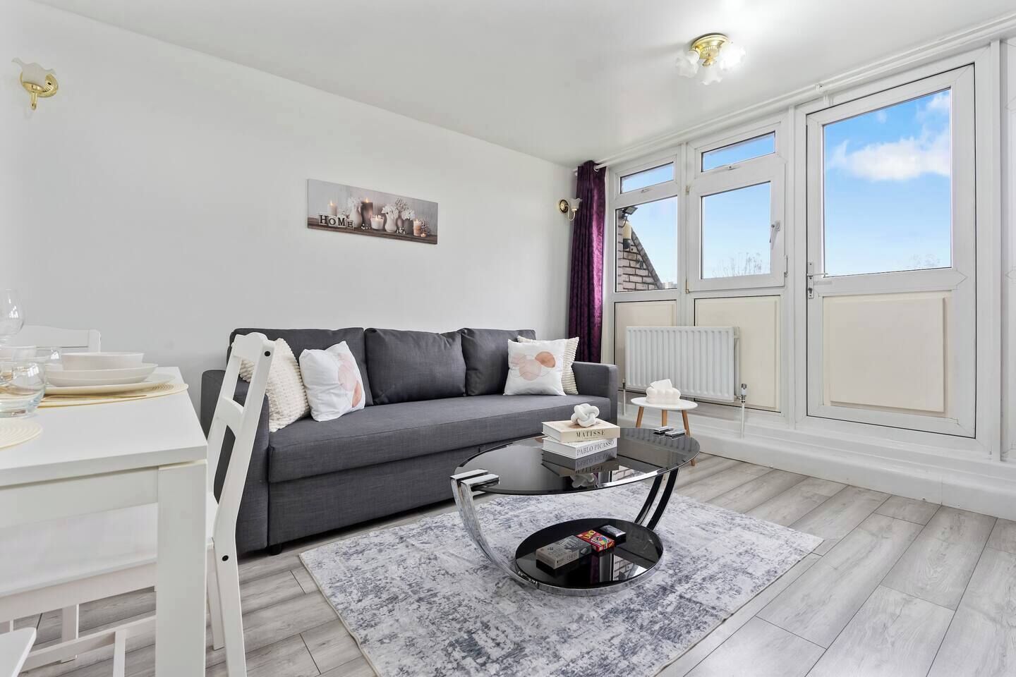 Un salon moderne avec un canapé gris, une table à manger blanche et une table basse noire. Une fenêtre avec des rideaux offre une vue sur la ville.