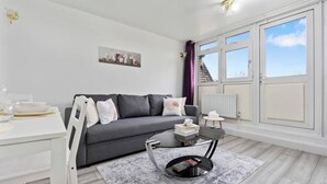 A modern living room with a grey sofa, a white dining table, and a black coffee table. There is a window with curtains and a view of the city.