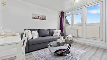Un salon moderne avec un canapé gris, une table à manger blanche et une table basse noire. Une fenêtre avec des rideaux offre une vue sur la ville.
