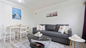 A modern living room with a grey sofa, white dining table, and chairs. There is a coffee table with books and a board game.