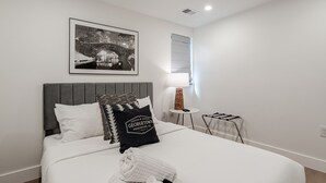 Een net opgemaakt bed met een grijs hoofdbord, een zwart-wit kussen en een lijst met een stadsbeeld.