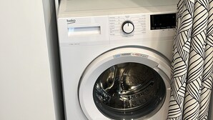 A compact laundry area with a front-loading washing machine, shelves above containing cleaning supplies, and a patterned curtain. - Standing apartment, heart of Bareges village, close to amenities, sleeps 1 to 8 (Barèges)