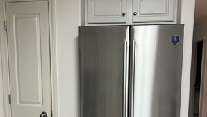 Frigorifero in acciaio inossidabile con due porte e un vano congelatore, posizionato in una cucina con mobili bianchi e pavimento piastrellato.