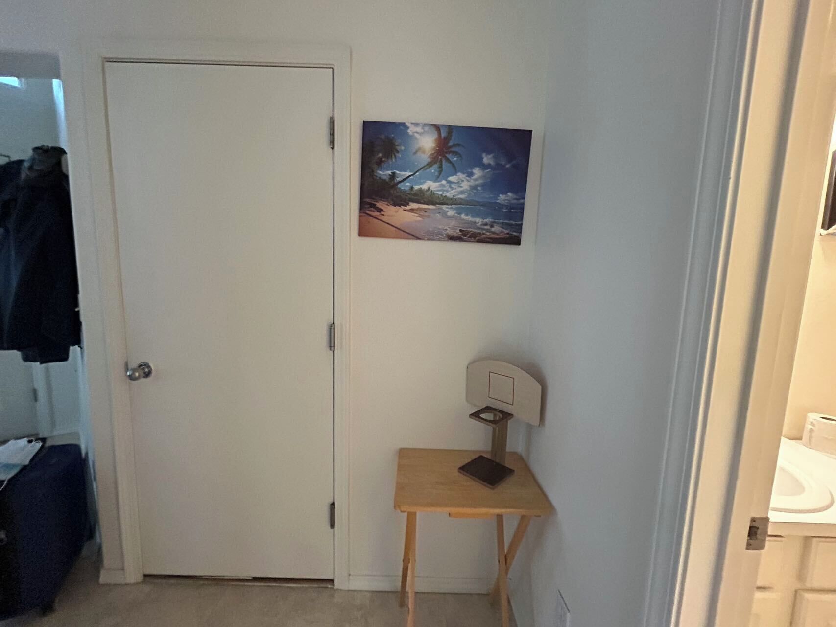 A room with a white door, a small wooden table with a laptop, and a painting of a beach scene on the wall.