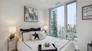 Un dormitorio moderno con una cama grande, vistas de la ciudad y un cuadro enmarcado de palmeras.