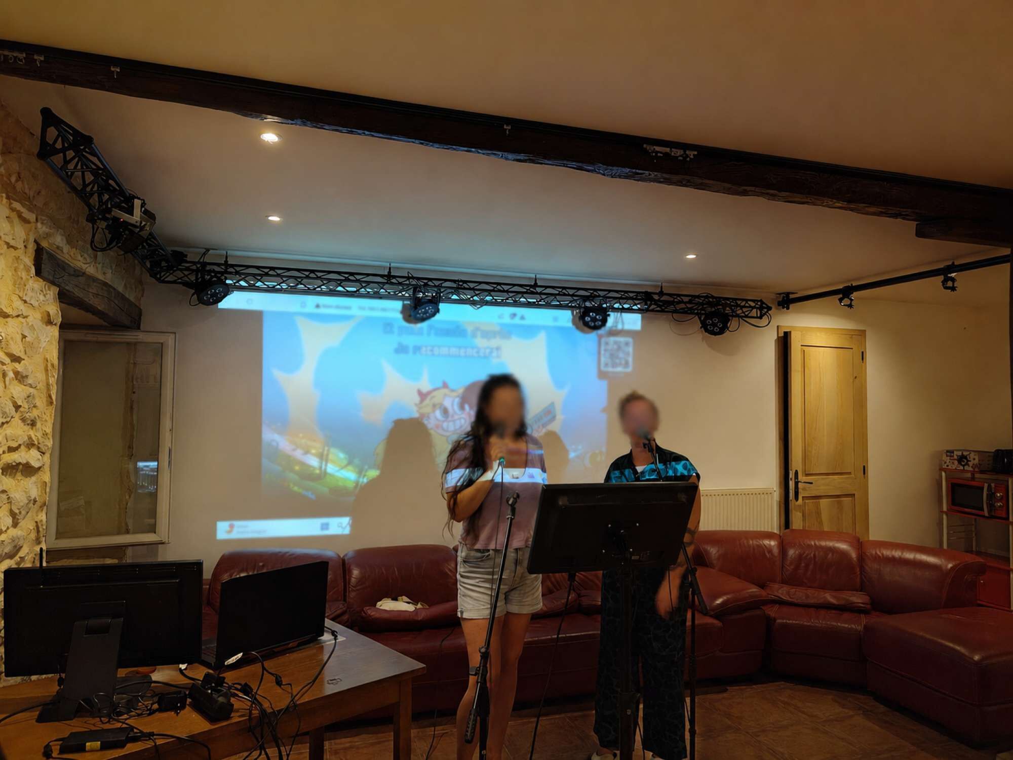 A room with a stone wall, a wooden ceiling, a red leather couch, a wooden table with a laptop, and a projection screen displaying a cartoon character.