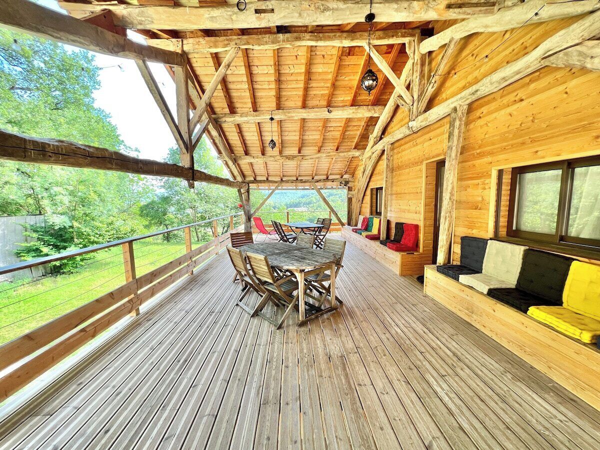 Een houten terras met een tafel, stoelen, een bank en een raam met uitzicht op groen.