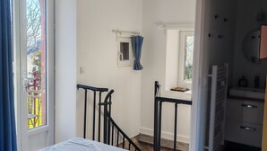 Un dormitorio con cama, una ventana con cortinas, una mesita con silla y un baño con espejo y lavamanos.