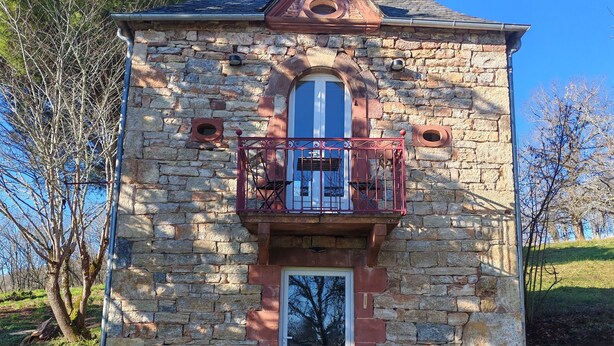 Un edificio de piedra con una puerta central, un pequeño balcón con barandilla y un camino de grava que conduce a la entrada.
