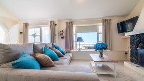 Un salón con un sofá beige, una mesa de centro blanca, un televisor de pantalla plana, una chimenea y vistas al mar a través de amplios ventanales.
