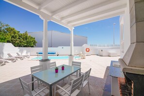 Un patio exterior con una mesa de vidrio, sillas blancas, una parrilla y una piscina al fondo.