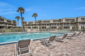 Une piscine entourée de chaises longues et de palmiers, le tout bordé par un immeuble doté de nombreux balcons.