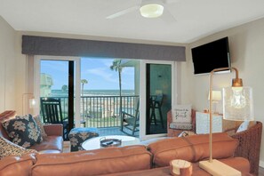 Sala de estar con sofá, sillones, una mesa de centro y televisor. Balcón con vista a la playa y palmeras.