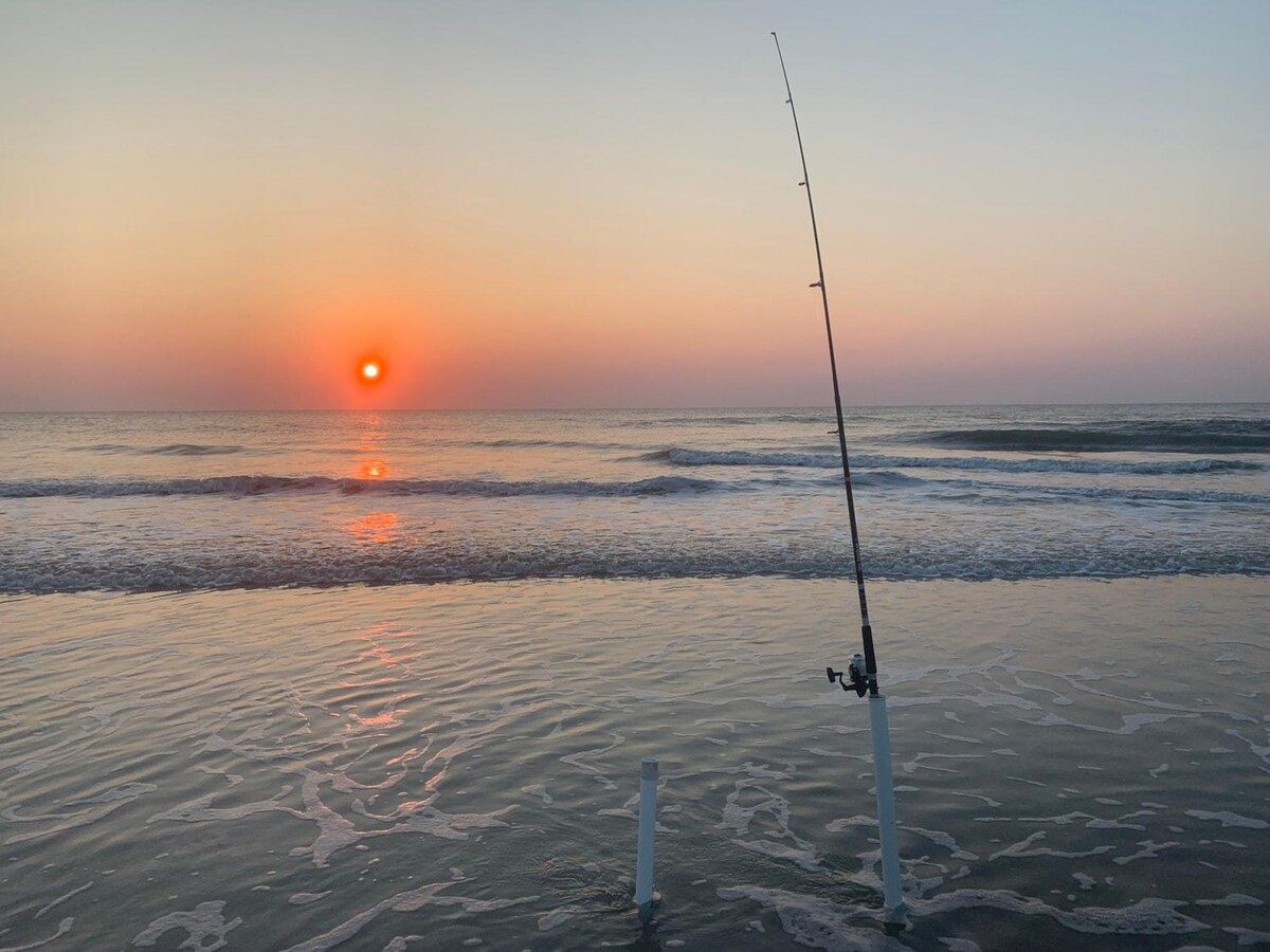 A fishing rod on a sandy beach with the sun setting in the background.