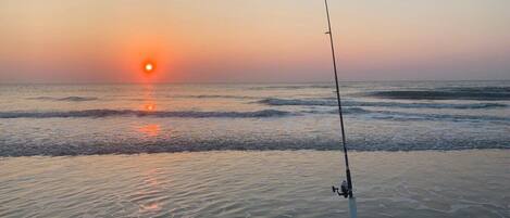 A fishing rod on a sandy beach with the sun setting in the background.