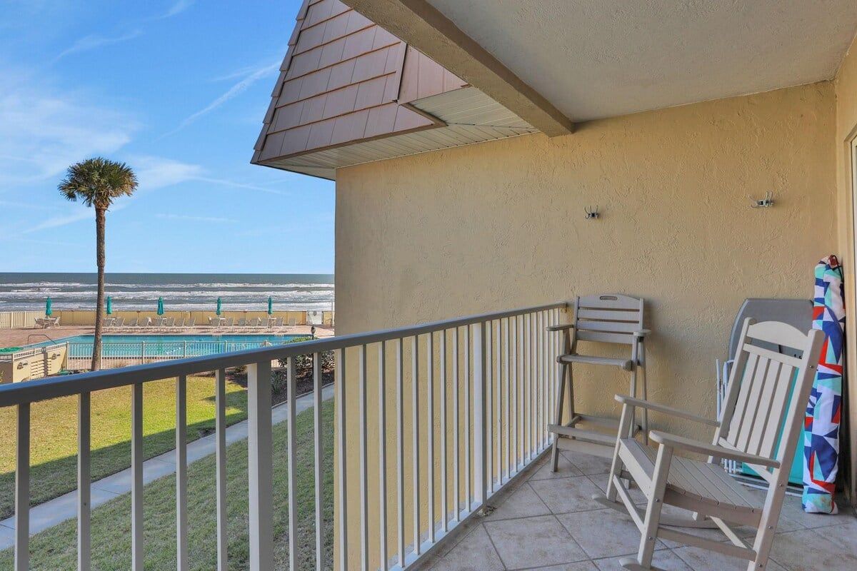 En balkon med udsigt til en swimmingpool og en palme, en stol og et lille bord.