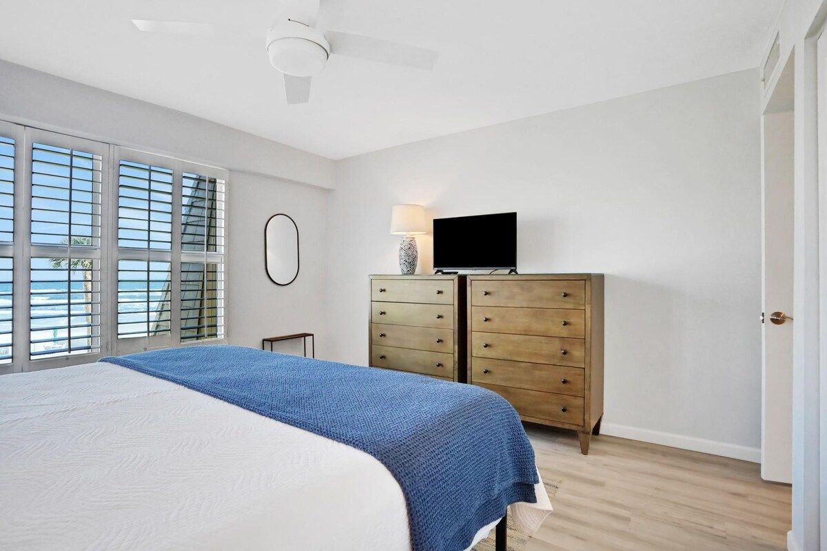 Une chambre à coucher comprenant un lit, une commode, un téléviseur et une fenêtre avec des stores.