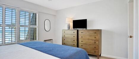 Une chambre à coucher comprenant un lit, une commode, un téléviseur et une fenêtre avec des stores.