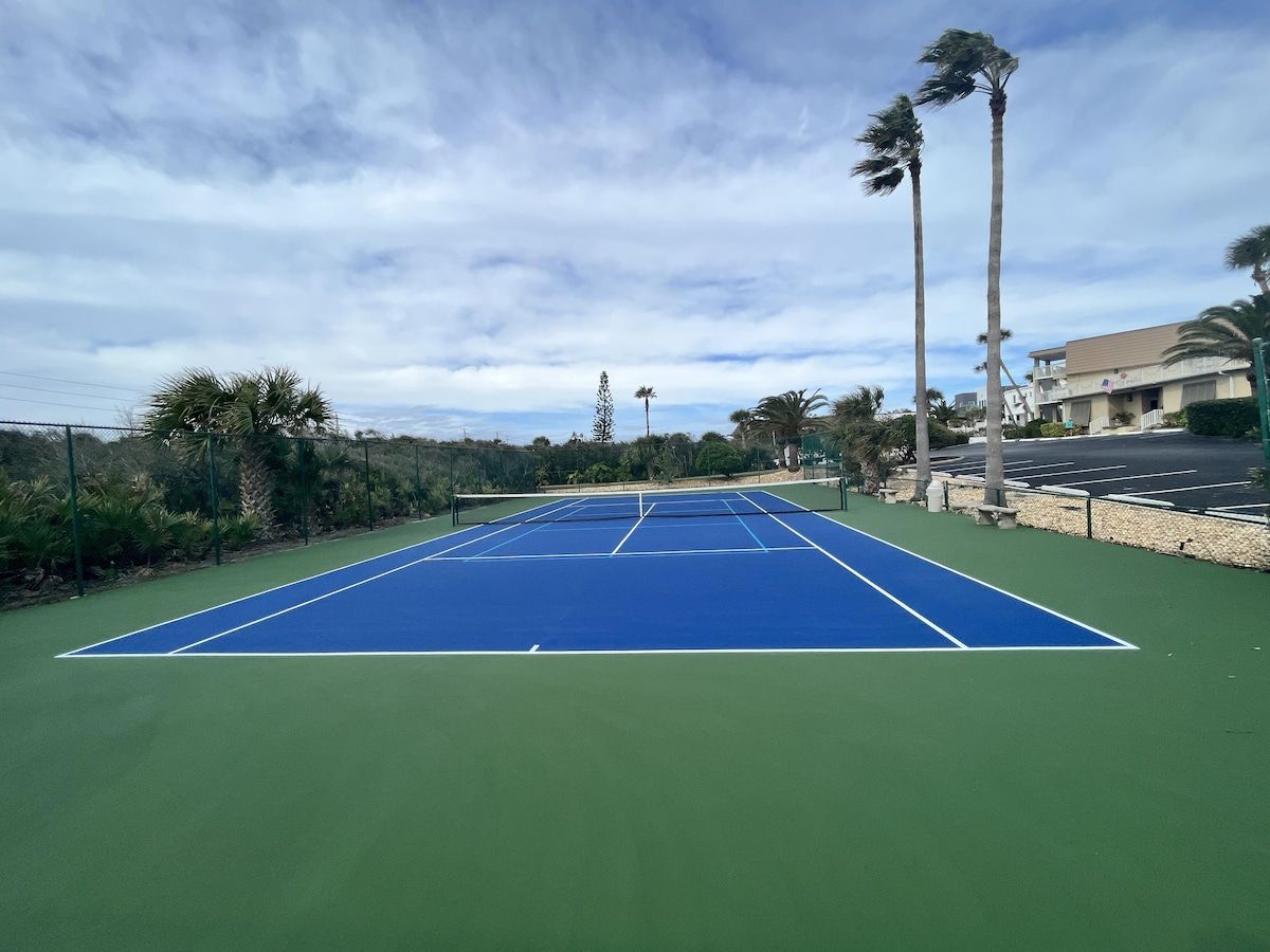 Un court de tennis avec une surface de jeu bleue et une zone périphérique verte, entouré de palmiers et avec un bâtiment visible en arrière-plan.
