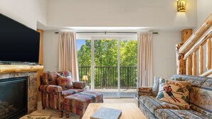 Un salon doté d’une cheminée, de meubles à motifs, d’un livre posé sur une table basse et d’un balcon offrant une vue sur les arbres.