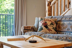 A room with a sofa featuring a patterned cushion, a wooden table with a book and a candle, and a view of greenery outside. - NEW Construction! Ski-In/Ski-Out Condo, fire place, Bunk room (Warren)